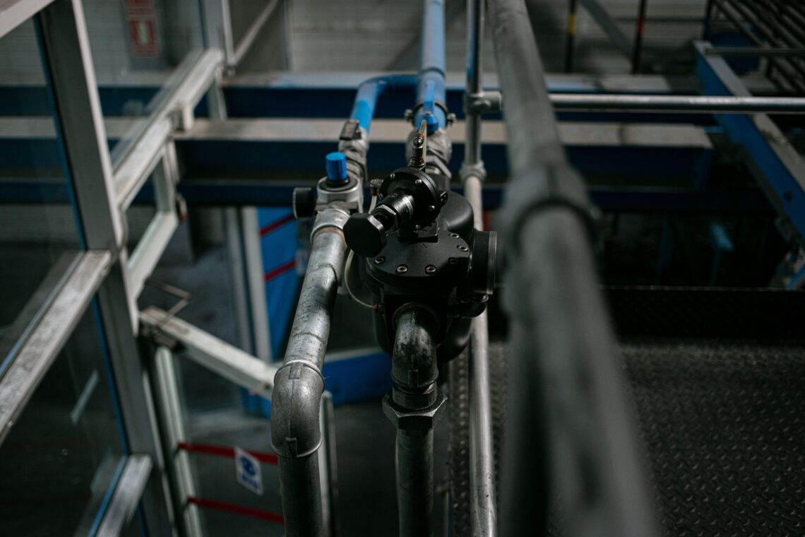 Galvanised process-water pipework inside a working industrial plant: a branch line carrying an in-line control body with its adjuster, tapped off the overhead main. This is the last meter of pipe — the point where a process's specified draw rate is either held, or left to follow whatever pressure the main happens to be carrying.